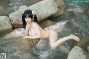 A woman in a white bikini sitting on a rock by the water.